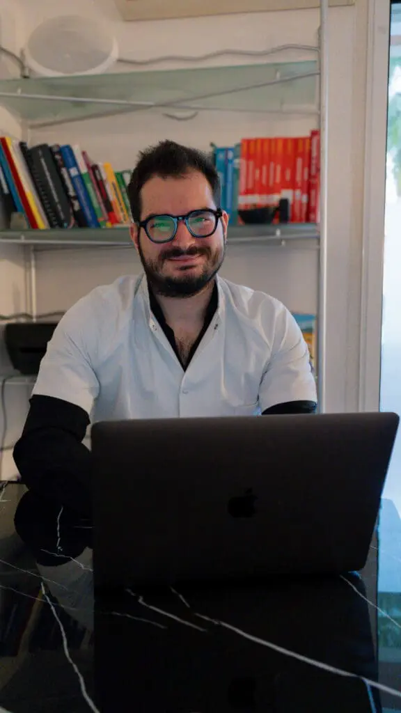 Ostéopathe à Nice souriant dans son cabinet, devant son ordinateur portable et sa bibliothèque médicale